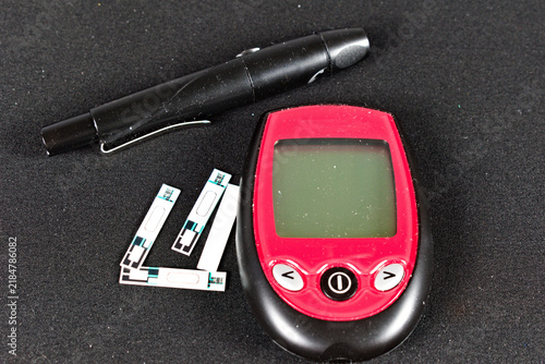фотографія A red and black glucose meter with a test strip inserted sits next to a black lancet pen on a textured dark surface