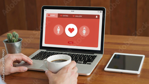 Laptop displaying dating app interface with heart icon and user profiles on a wooden desk with coffee