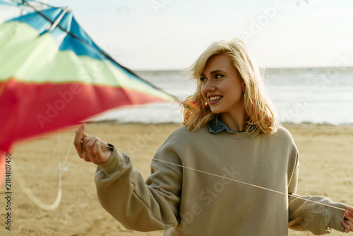 Smiling young adult Caucasian woman flying colorful kite on sandy beach outdoors. Image suited for tourism advertising, summer travel promotions, and outdoor recreation branding