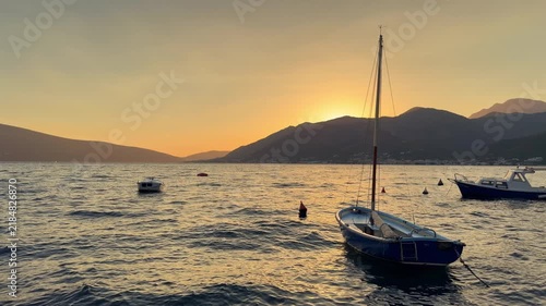 Small sailboat gently rocking on waves in the Adriatic Sea at sunset, with mountain coastline and other boats in the background