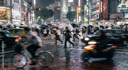 Busy Tokyo street scene at night with motion blur capturing pedestrians, vehicles, and neon signs reflecting on wet asphalt after rain