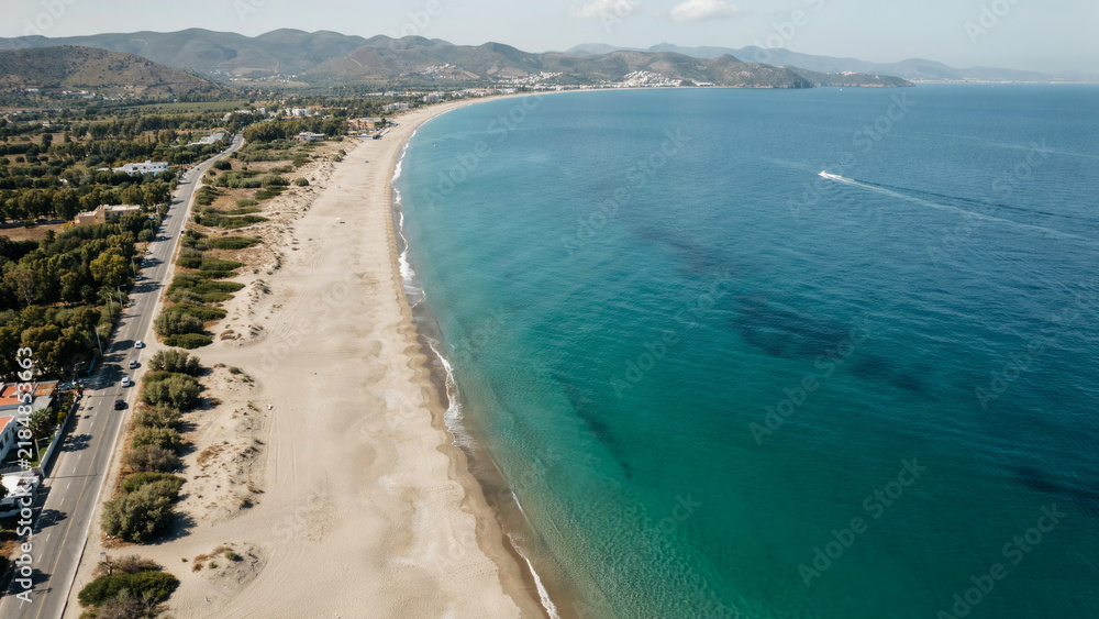 Obraz premium Panoramic view of long sandy beach next to turquoise sea and coastal road.