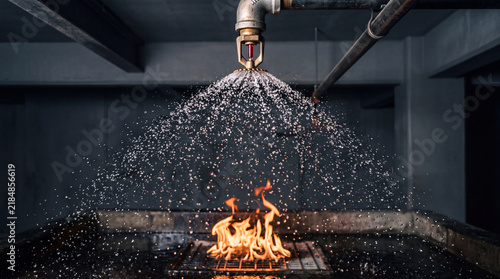 Industrial metal fire sprinkler head releasing water spray over burning bright orange flames in dark warehouse
