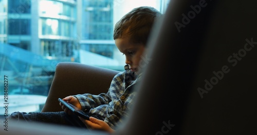 Принти на полотні Preschooler in plaid shirt engrossed in phone comfortably sitting on sofa in cafe waiting for parents