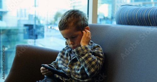 Plakat Boy preschooler intently watches smartphone screen while sitting on couch with hand on ear. Child absorbed posture highlights common scene of digital engagement