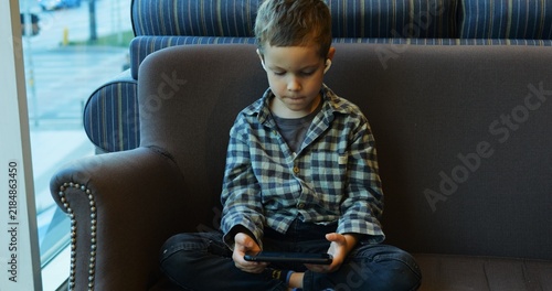 Fotografie Preschooler sits cross-legged on sofa in cafe engrossed in phone waiting for order