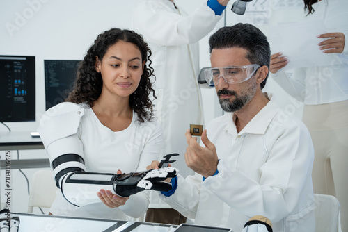 A group of engineers work together in a bright laboratory to test a robotic hand prototype and computer chip. A man holds a small microchip while colleagues examine mechanical prosthetic parts.
