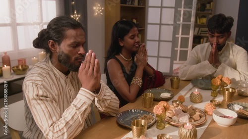 South Asian young adults sitting at festive table praying with folded hands. Visual perfectly serves cultural diversity campaigns, holiday storytelling, and religious festival marketing