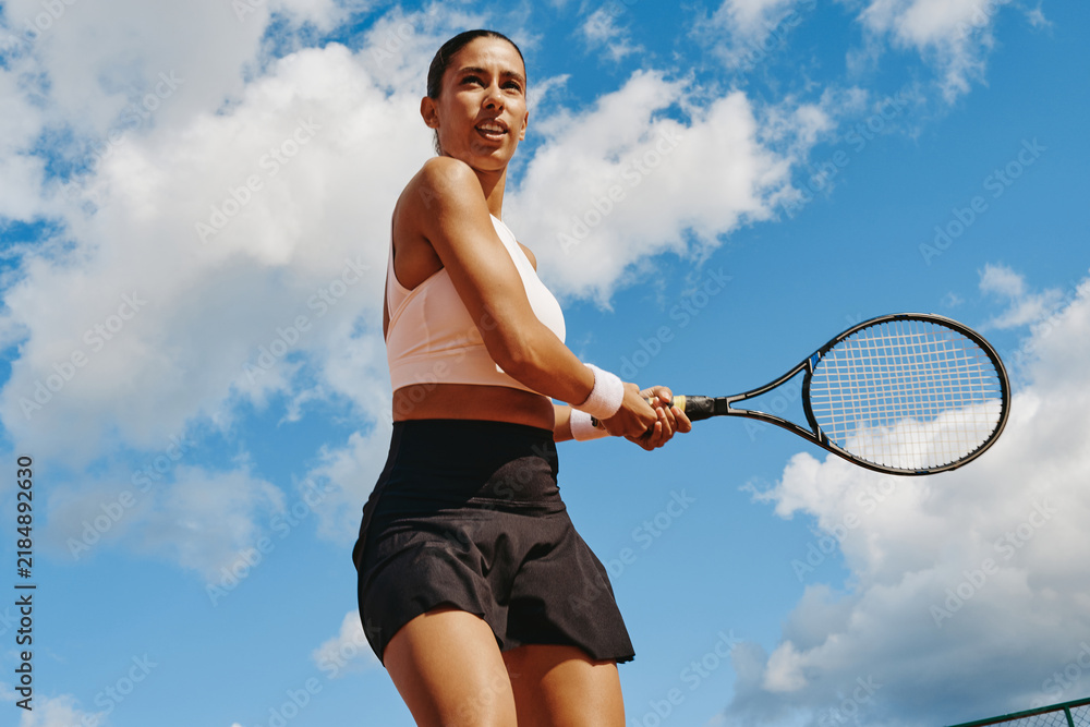 Obraz premium Multiethnic young adult woman holding racket ready to hit backhand during tennis match. Low angle view ideal for fitness advertising, active lifestyle campaigns and sports tournament promotion