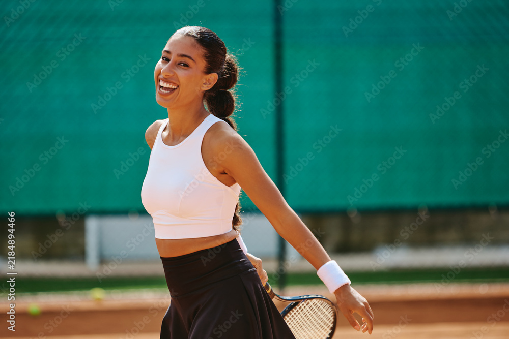 Obraz premium Multiethnic young adult woman holding racket and laughing on tennis court during training. Ideal for sports apparel advertising, fitness campaigns, recreation themes