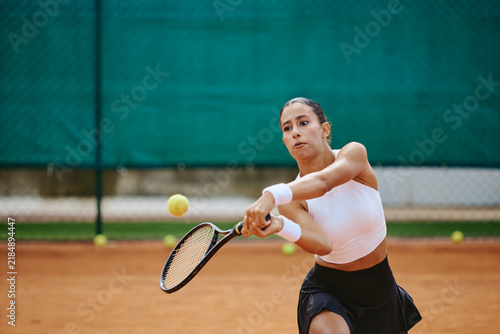 Bild auf Leinwand Young adult multiethnic woman playing tennis, hitting backhand stroke with racket on clay court