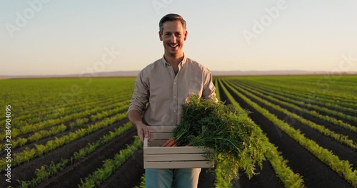Smiling farmer standing in a cultivated field holding a wooden box filled with freshly harvested organic carrots with green tops at sunset