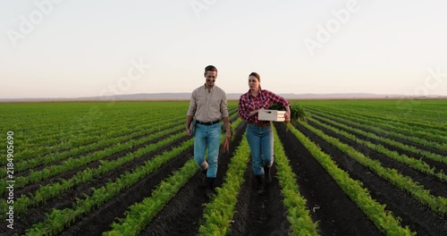 Two young farmers walking together through their agricultural field. The woman is carrying a wooden crate full of freshly harvested carrots