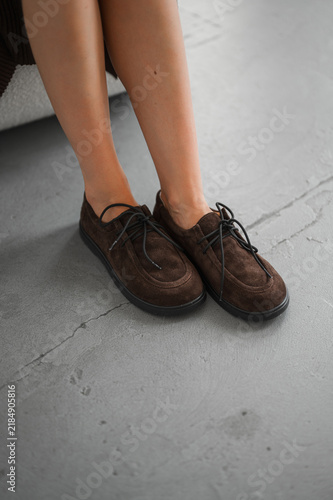 Close-up view of lower legs and feet wearing brown suede lace-up shoes on gray concrete floor with soft indoor lighting and casual fashion styling in relaxed pose