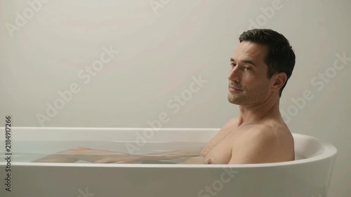 Man relaxing in white bathtub with eyes closed, meditating in bright minimalist bathroom. Peaceful wellness and self-care hygiene concept at home.