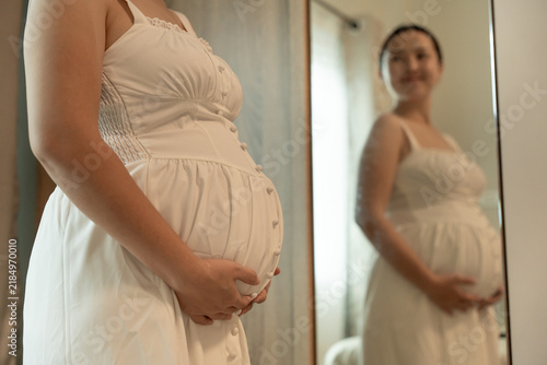 Close-up of a pregnant woman's belly in a white button-front dress, her smiling reflection visible in the mirror behind.