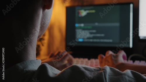 Over the shoulder view of a teenager typing code on a computer at a desk in a dark room.