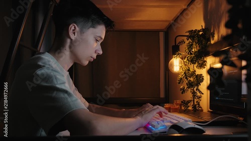 Side view of a focused teenage boy typing on a backlit keyboard while coding on a computer in a cozy, dark bedroom at night.