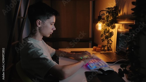 Focused teenage boy coding on a computer with a glowing keyboard in a dark room under warm light at night.