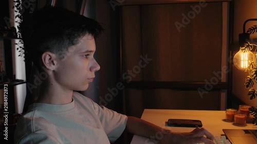 Side view of a teenage boy working on a computer at his desk in a dimly lit room at night.