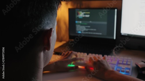 Rear view of a teenage boy writing code on a backlit RGB keyboard at a computer desk in a dark room.