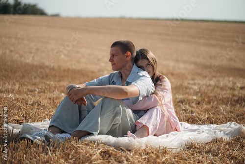 A couple sits together on a blanket in a wheat field while the woman leans against the man. The image represents love, companionship, and shared relaxation.