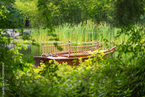 Canvas Print A wooden pleasure boat with a railed deck floats on a pond, with ducks resting aboard, framed by green leaves and tall reeds behind