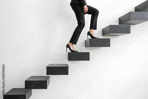 A woman wearing a black business suit and heels walking up a set of floating black stairs against a plain white background, for success