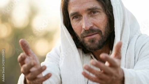 Jesus reaching out his open hands to the viewer in warm light. Close up of Christ in a white hooded robe with a soft bokeh background. Faith, invitation and salvation