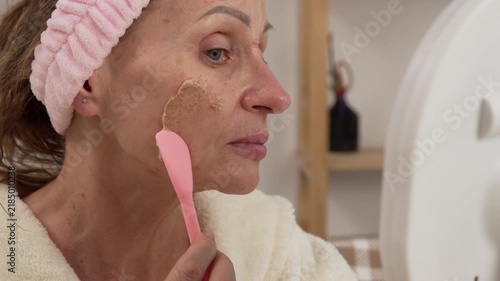 woman in a bathrobe applying a scrub to her face with a spatula, enjoying her skincare routine, creating a relaxing atmosphere at home during morning rituals.