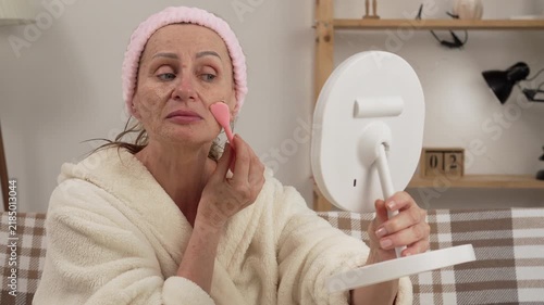 woman in a bathrobe applying a scrub to her face with a spatula, enjoying her skincare routine, creating a relaxing atmosphere at home during her morning