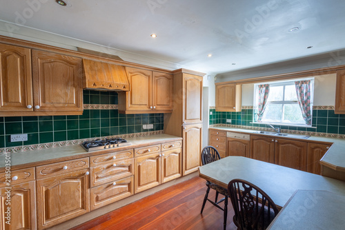 Wide view of a traditional 1990s fitted kitchen with wooden cabinets dark green tiled splashback gas hob sink window worktops table chairs and hardwood flooring in an older home
