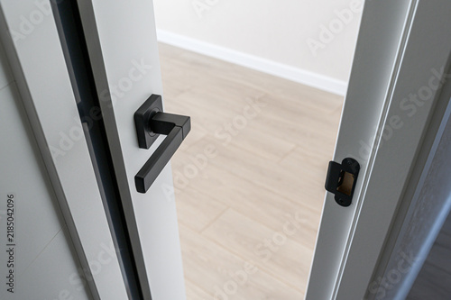 Matte black geometric door handle on white door with black trim, visible latch plate, clean angular lines, minimalist design, light wood floor, neutral interior space
