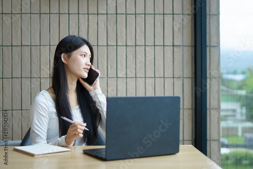 Young Asian businesswoman multitasking talking on the phone while note and using laptop at a modern office desk, smiling confidently as she works on remote tasks and virtual meetings