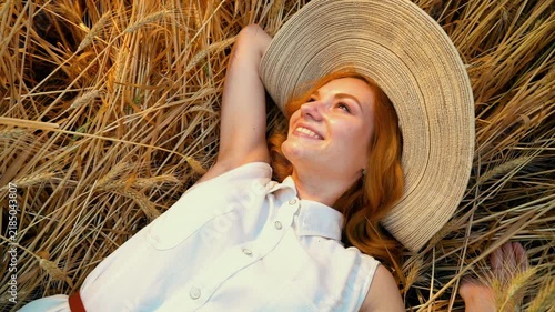 Cheerful young ginger girl in straw hat looking away and smiling while resting on ground. Beautiful female model enjoying summer relaxation
