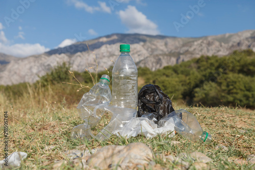 Plastic bottles lie scattered in dry grass. Black trash bag rests nearby, half-buried. Distant mountains frame the polluted scene. Blue sky with soft clouds adds contrast