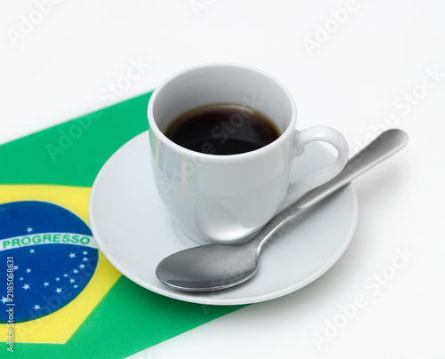 White cup of coffee on saucer next to the Brazilian flag on white background