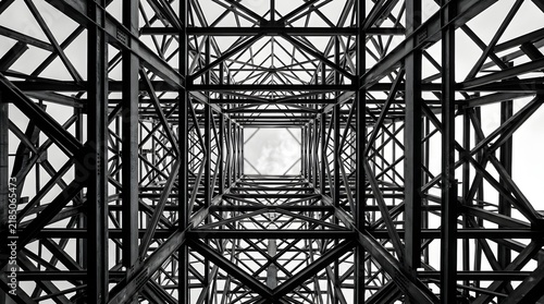 Looking upward through an intricate lattice of steel beams that converge into a square opening against a pale sky  emphasizing industrial geometry in monochrome