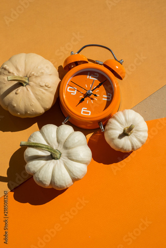 Orange alarm clock with three white decorative pumpkins on two tone beige and orange background for autumn season and daylight saving time concept