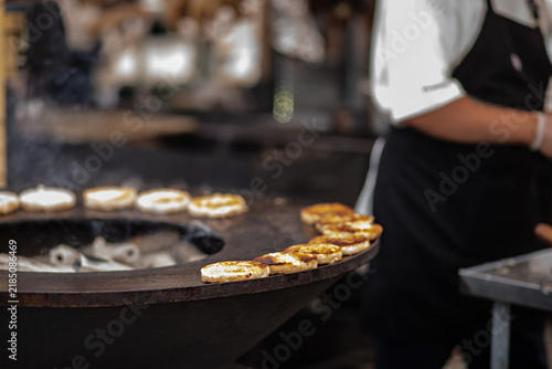 Chef grilling meat patties on round charcoal grill, brushing sauce, smoke rising, outdoor street food market cooking.