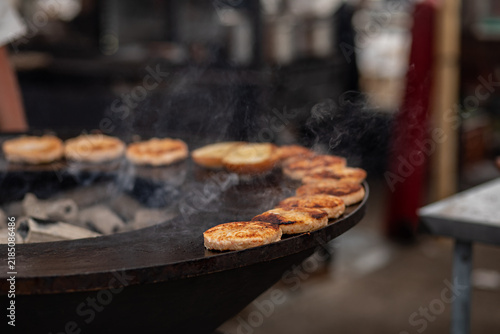 Chef grilling meat patties on round charcoal grill, brushing sauce, smoke rising, outdoor street food market cooking.