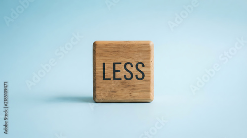 A single square wooden block with the word less engraved in black capital letters, centered on a soft light blue background, representing minimalism, simplicity, and the concept of doing less