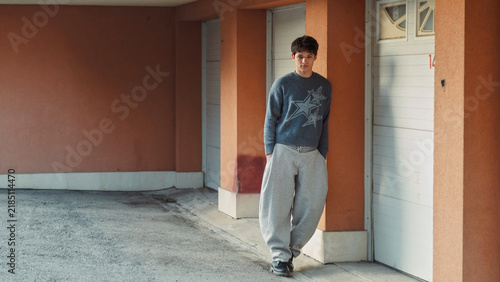 Teenage boy in grey sweatpants leans against a terracotta orange wall beside a doorway, a warm colour street style portrait with strong architecture for youth fashion brands