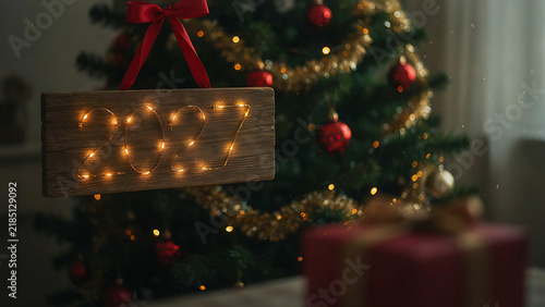Hanging wooden board with illuminated 2027 fairy lights and red ribbon before christmas tree, created with generative ai.