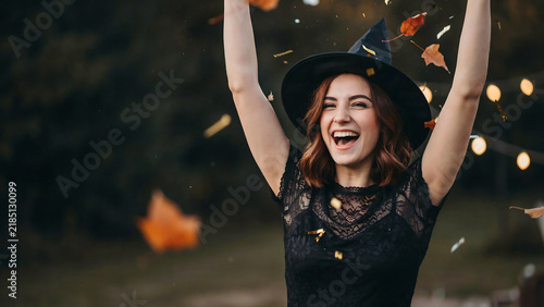 Laughing woman in witch hat celebrating with gold confetti and falling leaves near blurred child, created with generative ai.