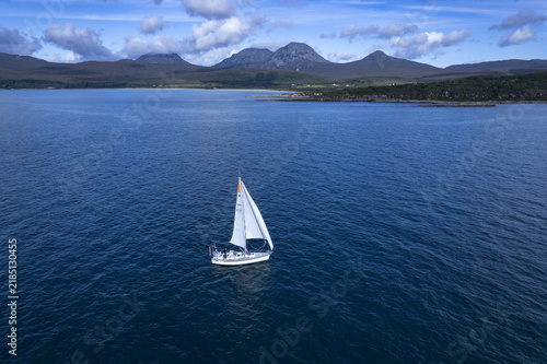 Isle of Jura, Scotland, United Kingdom - 24 August 2022: Aerial view of a sailboat on deep blue waters near the Isle of Jura coast with the Paps of Jura mountains in the background.
