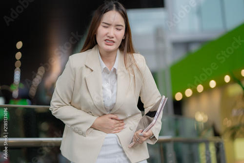 Stomach pain concept of a young businesswoman holding her abdomen with discomfort while carrying a tablet at work. stomach pain, abdominal pain, woman, discomfort, workplace