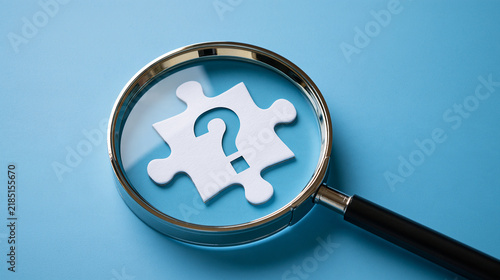 Magnifying glass examining a white puzzle piece with a question mark on blue background. Problem solving concept for business strategy presentations.