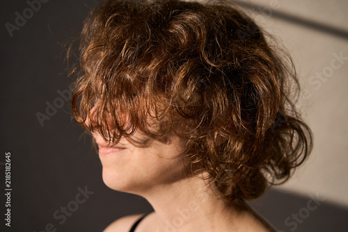 Young Woman With Curly Hair In Side Profile Smiling In Warm Indoor Sunlight Near Plain Wall.