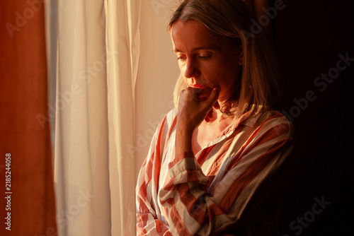 Bild auf Leinwand Pensive middle aged woman supports chin with hand beside glowing curtains